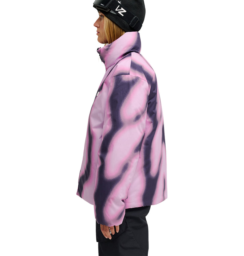 A person in a black helmet and black pants is wearing the DC Shoes Aura Puffer Jacket—a pink and purple tie-dye waterproof jacket—standing in profile against a white background.