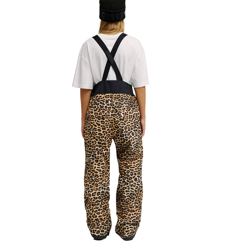 A person stands facing away, wearing a white t-shirt, black suspenders attached to the DC Shoes Valiant Bib Pants—with their distinctive leopard print offering breathable protection—black shoes, and a black beanie hat.