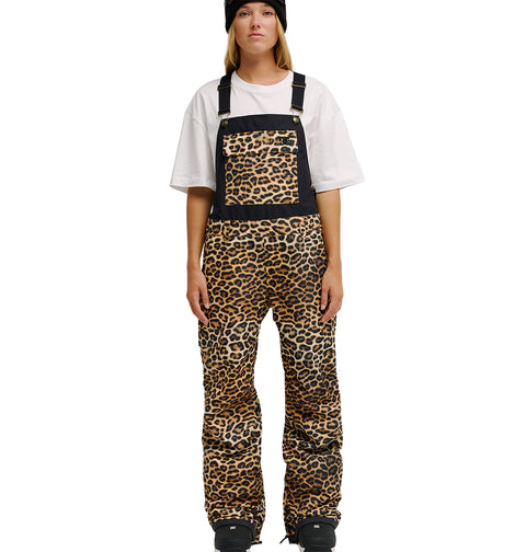 A person wearing the DC Shoes Valiant Bib Pants stands facing forward, pairing the leopard print overalls—with breathable protection, a front pocket, and black accents at the top—with a black beanie and white oversized t-shirt.