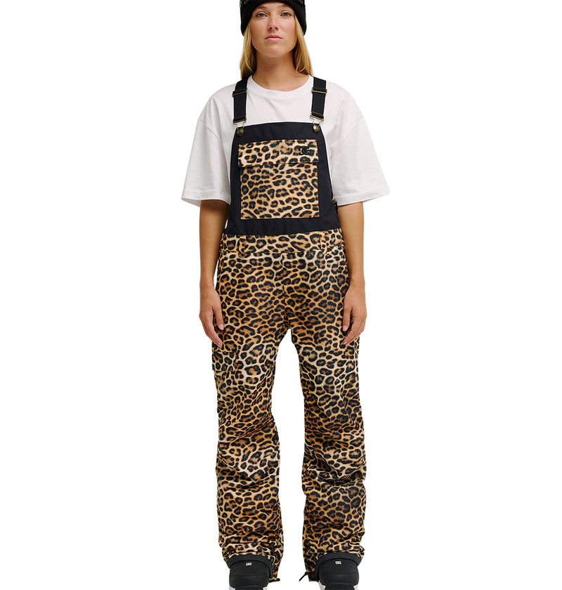 A person wearing the DC Shoes Valiant Bib Pants stands facing forward, pairing the leopard print overalls—with breathable protection, a front pocket, and black accents at the top—with a black beanie and white oversized t-shirt.