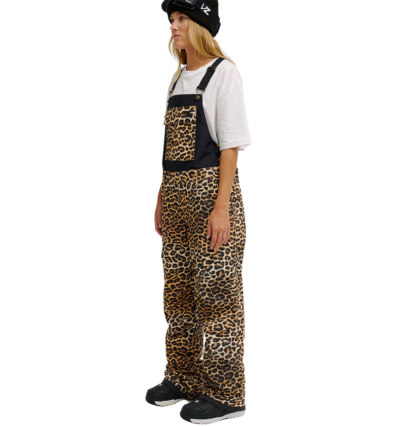 A person stands sideways against a white background, wearing a black hat, straight blonde hair, and black sneakers. They are dressed in DC Shoes Valiant Bib Pants featuring a leopard-print design with black straps over a white T-shirt, offering breathable protection for all-day comfort.
