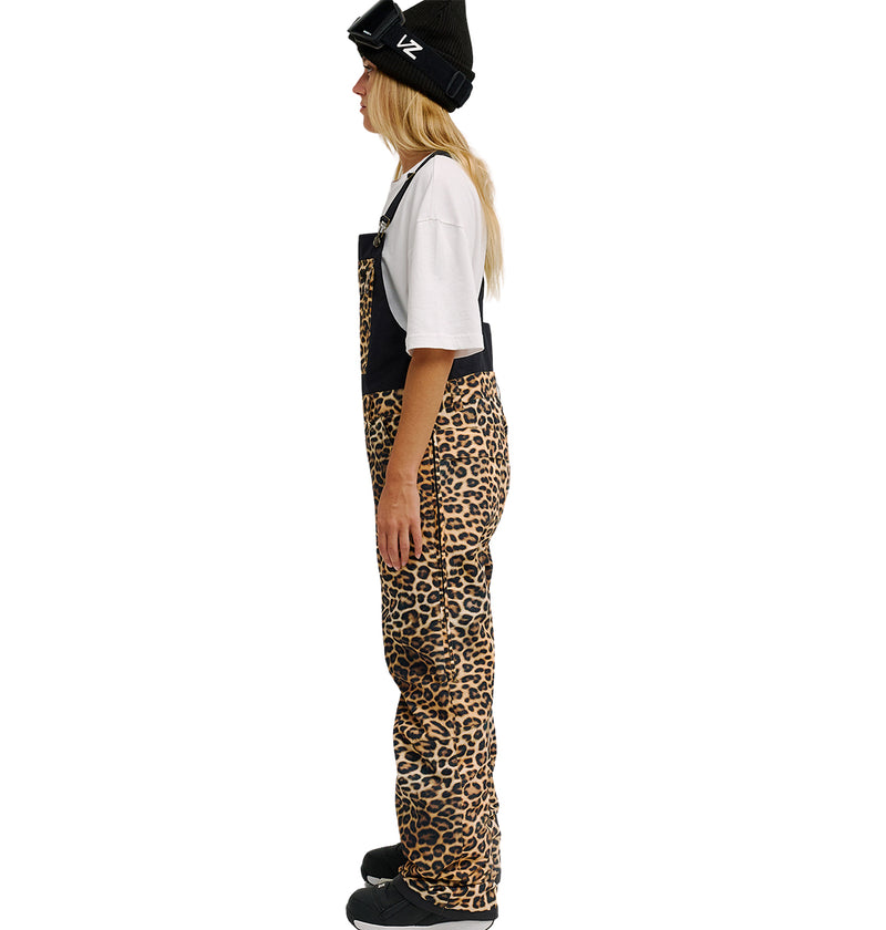 Wearing DC Shoes’ Valiant Bib Pants—leopard print overalls designed for breathable protection—paired with a black beanie, goggles, white t-shirt, and black boots, a person in profile stands against a plain white background.