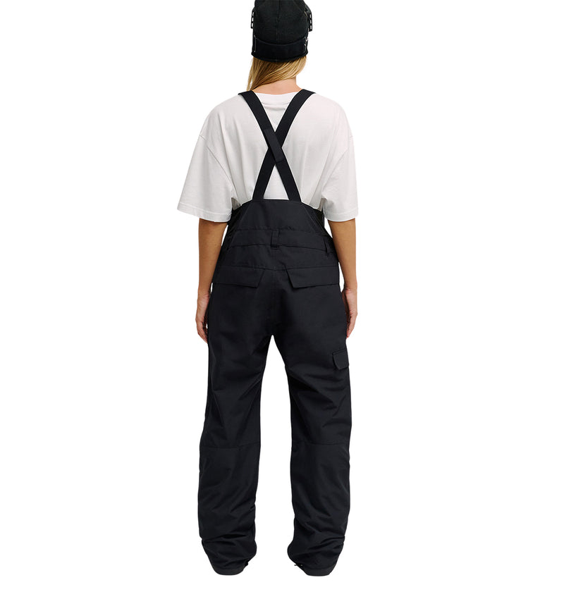 A person stands with their back to the camera against a plain white background, wearing a white t-shirt, black cap, and DC Shoes Valiant Bib Pants featuring crossed straps designed for breathable protection.