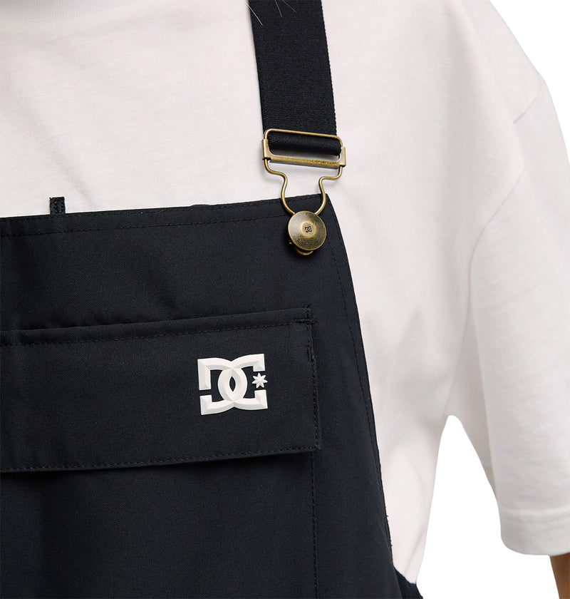 A person wears the Valiant Bib Pants by DC Shoes—black overalls with a front pocket, gold buckle, and a signature logo featuring interlocking DC letters with a star—paired with a white T-shirt. Designed for breathable protection and comfort during everyday wear.
