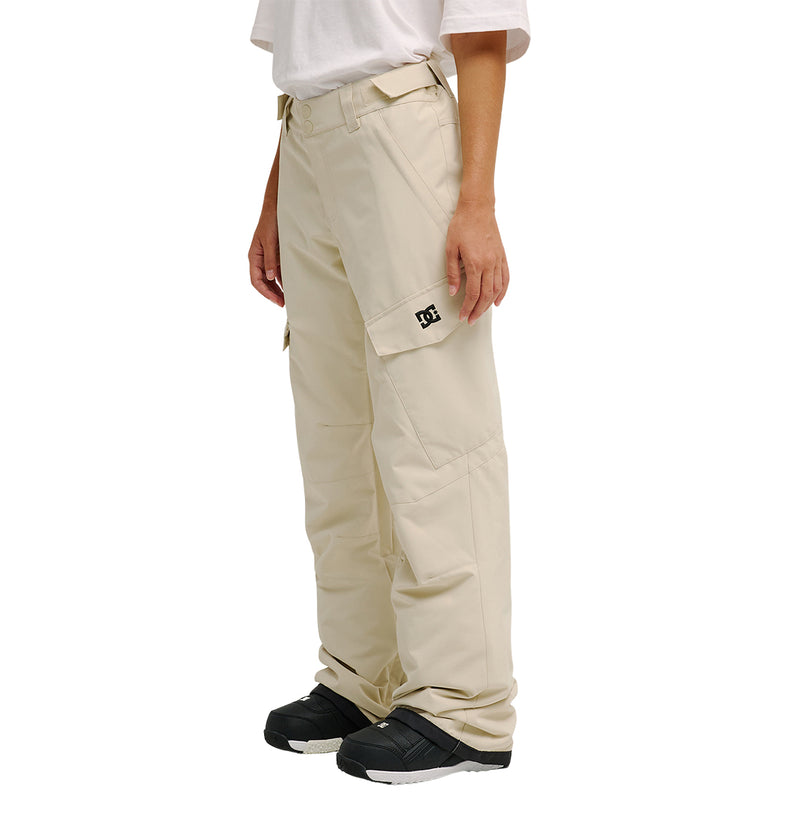 A person is shown from the waist down, wearing DC Shoes Nonchalant Pants—loose-fitting beige cargo pants featuring a small black logo on the pocket, designed for superior snow performance and dependable waterproofing. They are paired with a white t-shirt and black sneakers, standing against a plain white background.