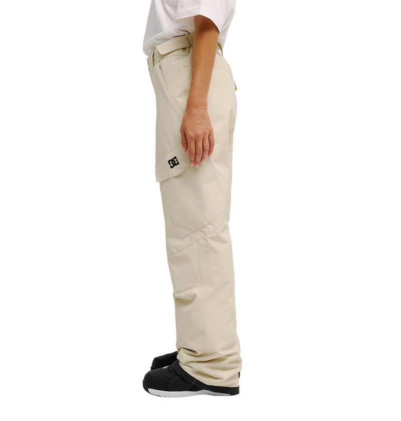 A person stands sideways against a plain white background wearing the Nonchalant Pants by DC Shoes—beige, waterproof, and insulated cargo pants featuring a side pocket with a small black DC logo—paired with a white t-shirt and black shoes.