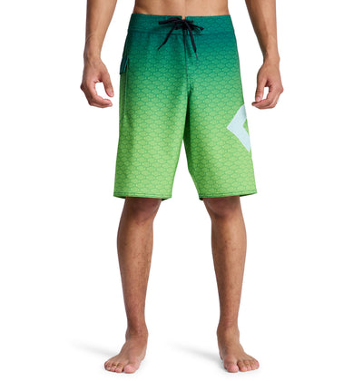 A man models Reebok's Lanai 21 board shorts—green gradient with a small geometric pattern, black drawstring, and a white angular design on the left leg—standing barefoot against a white background.