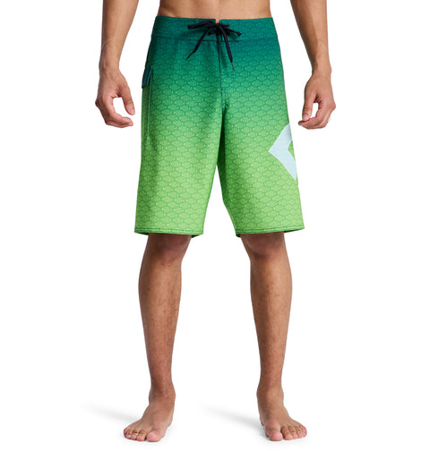 A man models Reebok's Lanai 21 board shorts—green gradient with a small geometric pattern, black drawstring, and a white angular design on the left leg—standing barefoot against a white background.