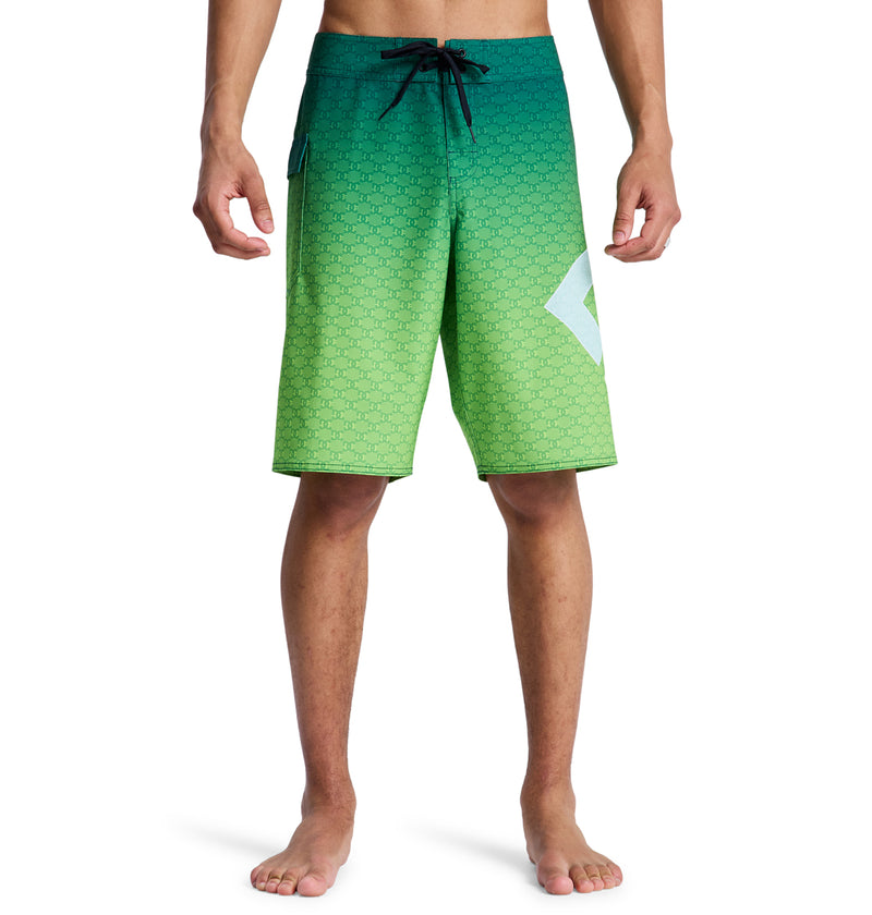 A man models Reebok's Lanai 21 board shorts—green gradient with a small geometric pattern, black drawstring, and a white angular design on the left leg—standing barefoot against a white background.