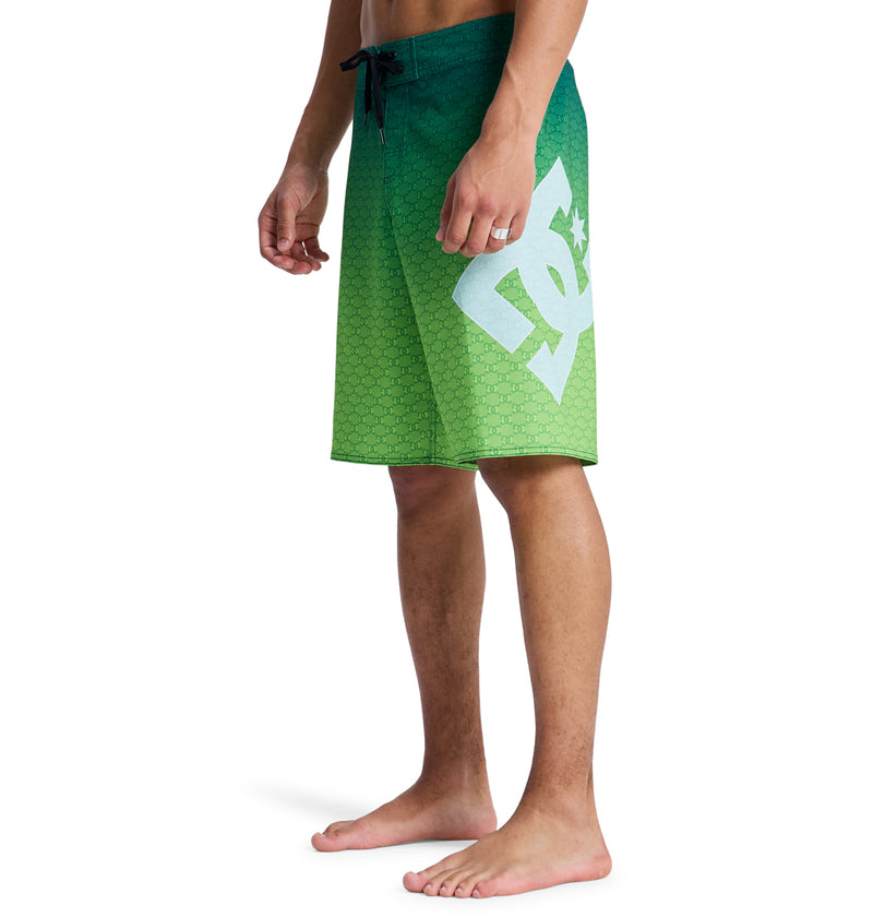 A man stands barefoot wearing Reebok Lanai 21 green gradient board shorts with a large white logo on the left thigh, hands relaxed by his sides, against a plain white background.
