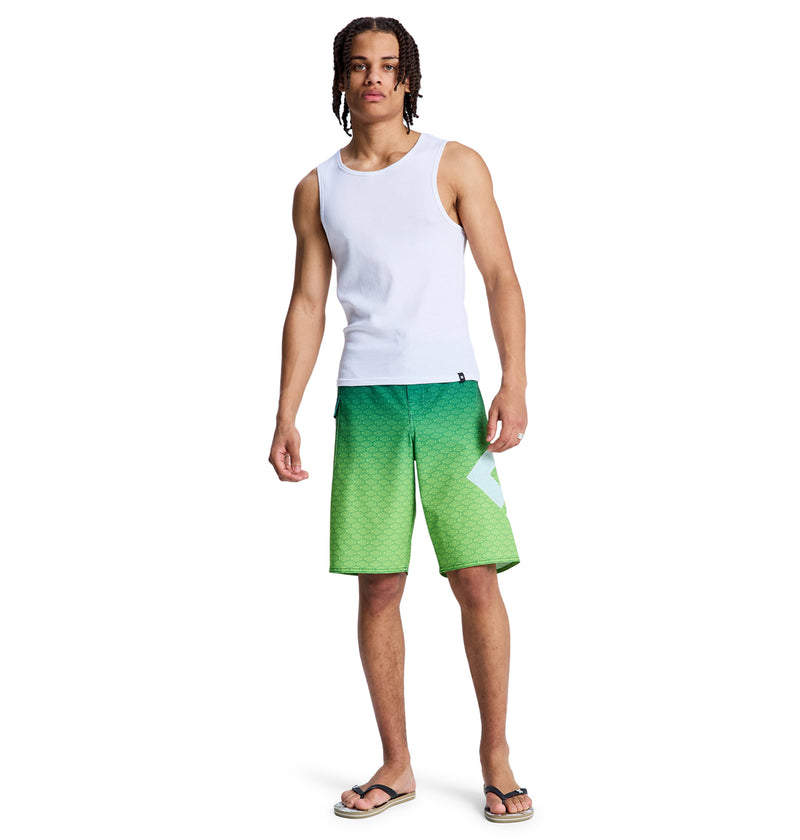 A young man with medium skin tone, wearing Reebok's Lanai 21 green patterned shorts and a white tank top with black flip-flops, stands facing forward against a plain white background.
