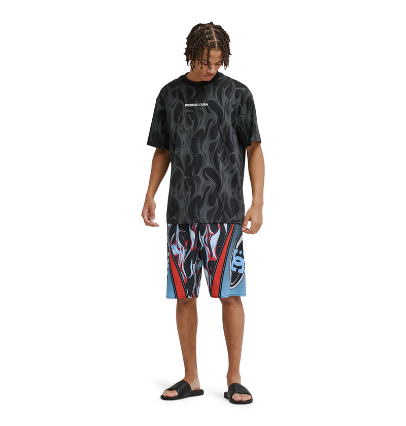 A person stands against a white background wearing the Reebok Sylem 21 black flame-patterned t-shirt, colorful flame-print shorts with red, white, and blue designs, and black slide sandals.