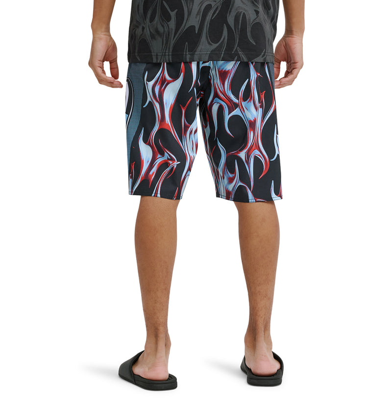 A person stands facing away, wearing Reebok Sylem 21 black and red flame-patterned shorts, a dark shirt, and black slide sandals, shown from the knees down against a white background.