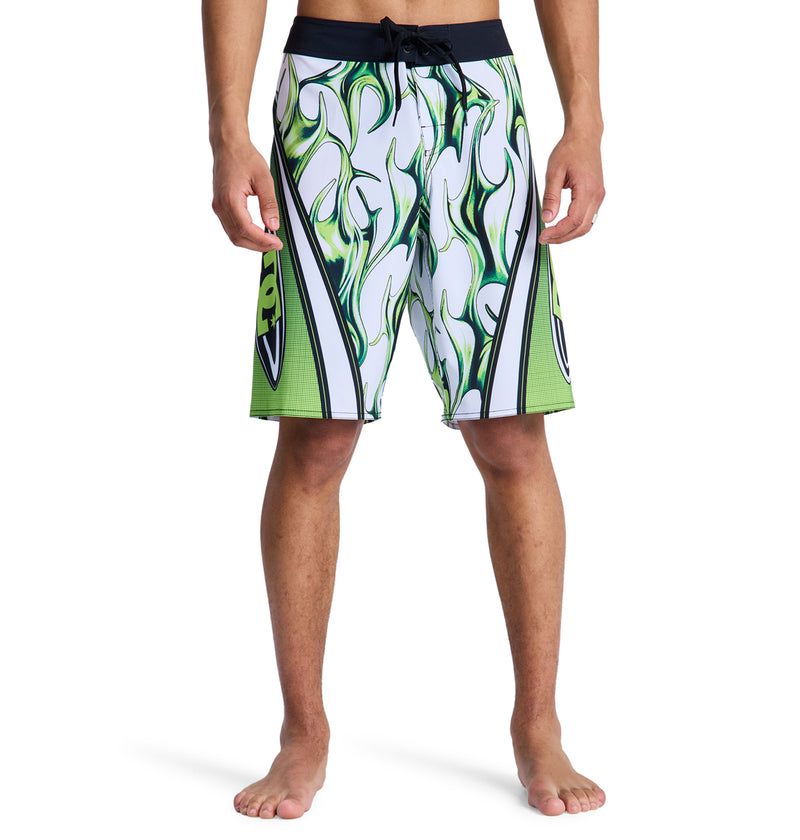 A person is seen barefoot, wearing Reebok Sylem 21 white board shorts featuring green and black abstract flame designs. The image is cropped at the waist and below the knees.