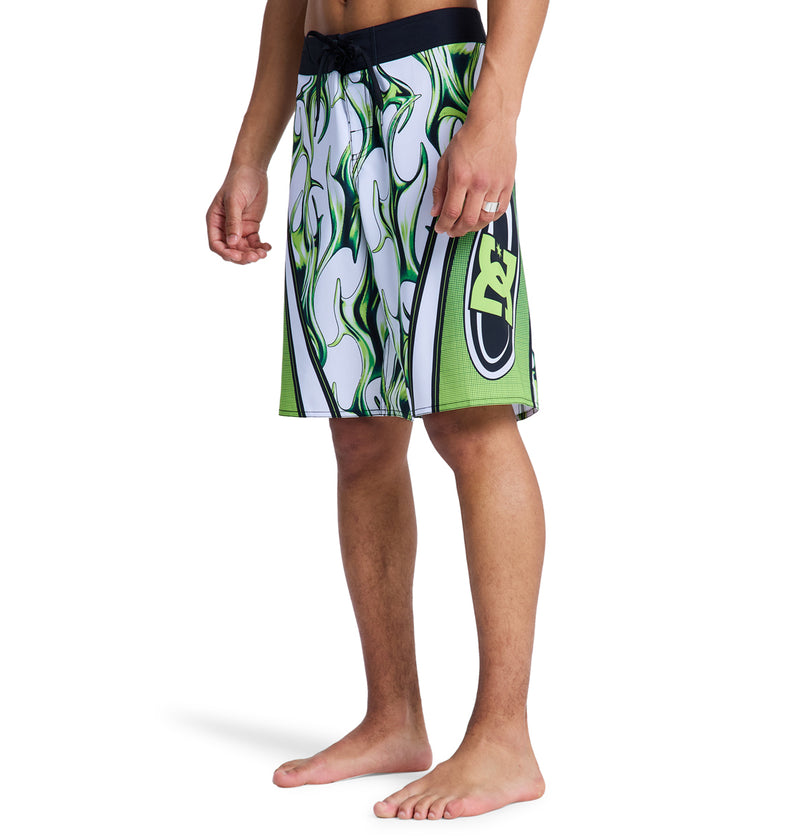 A person is barefoot, wearing Reebok Sylem 21 white knee-length swim trunks with green and black flame patterns and a large side logo. The upper body is not visible.
