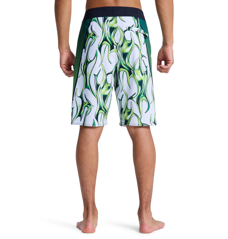 A person stands barefoot, facing away, wearing Reebok Sylem 21 board shorts with a black waistband and side panels, featuring a bold white and green patterned design with wavy green flames.