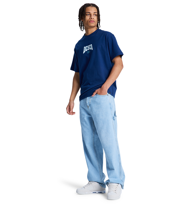 A person wears a navy DC Shoes t-shirt, white sneakers, and DC Shoes Men's Baggy Denim Carpenter Pant in a wide-leg fit, standing against a plain white background.