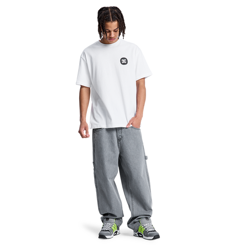 A young person with braided hair, wearing a white DC Shoes t-shirt, Men's Baggy Denim Carpenter Pant by DC Shoes, and black and green sneakers stands looking down with one hand in their pocket against a plain white background.