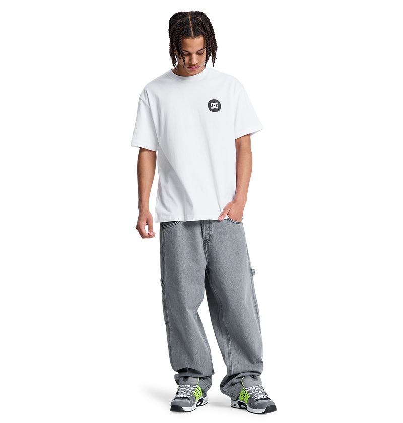 A young person with braided hair, wearing a white DC Shoes t-shirt, Men's Baggy Denim Carpenter Pant by DC Shoes, and black and green sneakers stands looking down with one hand in their pocket against a plain white background.