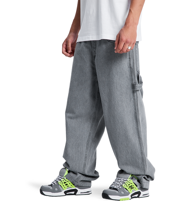 A person wears a white T-shirt, gray sneakers with white, black, and neon green accents, and DC Shoes Men's Baggy Denim Carpenter Pant—a wide-leg fit featuring heavyweight denim and side pocket detail that covers part of the sneakers.
