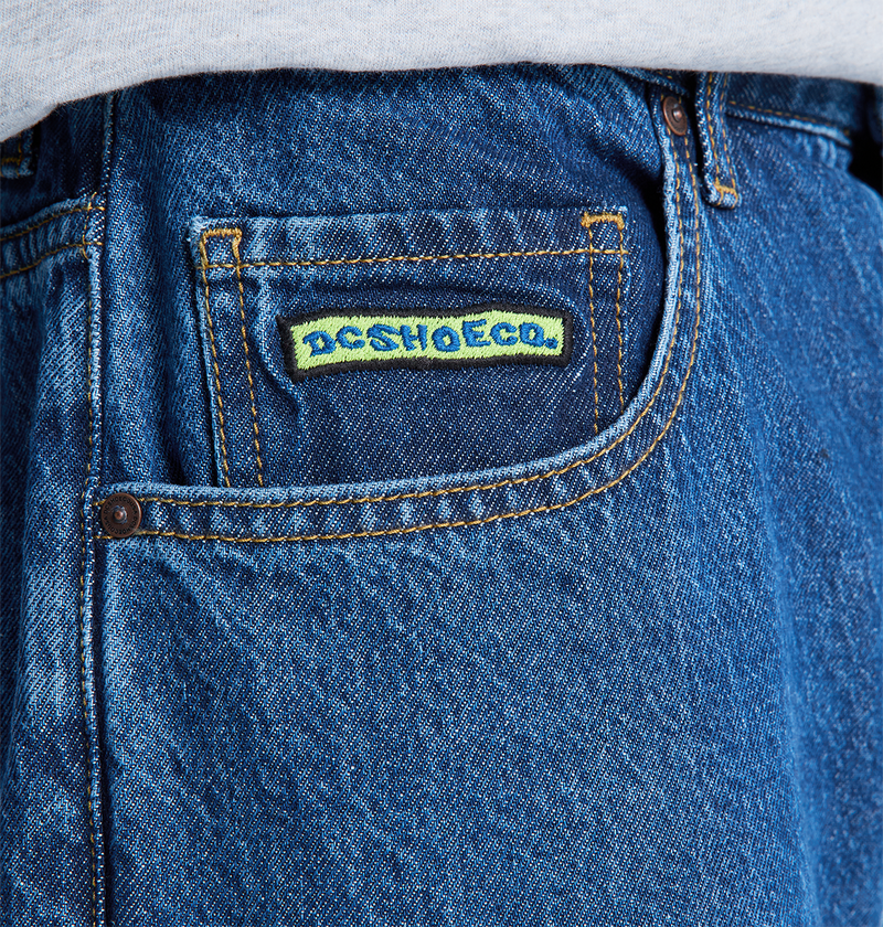 Close-up of the front pocket on DC Shoes Men's Baggy Denim Pant in blue heavyweight denim, featuring a yellow and blue “DC SHOESCO.” label above the pocket, with a white shirt partially visible at the top.
