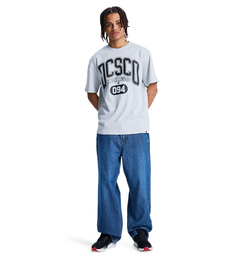 A person with braided hair wears a light gray t-shirt with DISCO and 094, Men's Baggy Denim Pant by DC Shoes in heavyweight denim, and black DC Shoes sneakers, standing against a plain white background.
