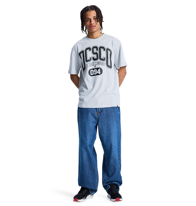 A person with braided hair wears a light gray t-shirt with DISCO and 094, Men's Baggy Denim Pant by DC Shoes in heavyweight denim, and black DC Shoes sneakers, standing against a plain white background.