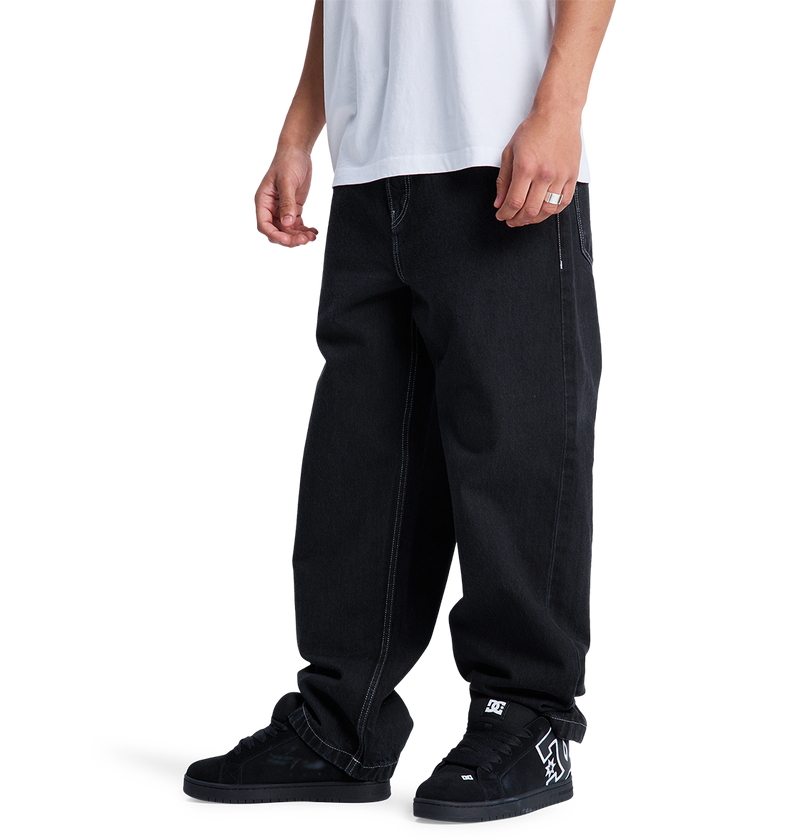 A model wears DC Shoes Men's Baggy Denim Pant and black DC skate shoes with a visible logo, paired with a white T-shirt. Cropped at the neck, the image highlights the relaxed, lower-body skate style against a white background.