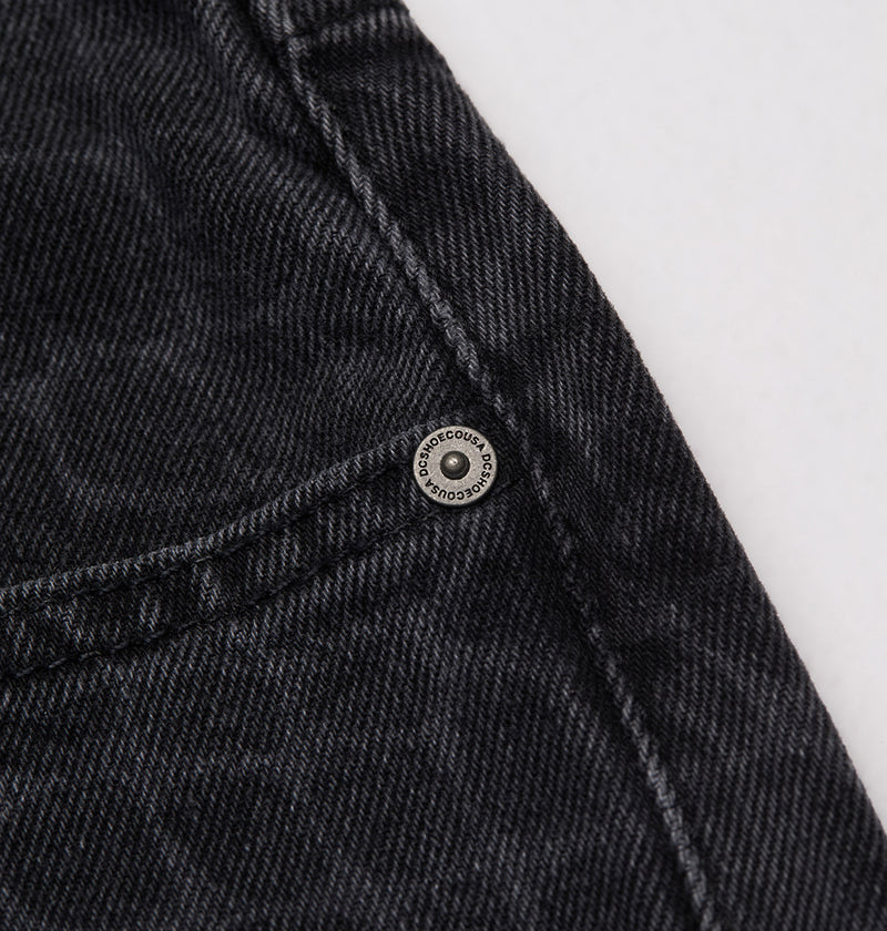 Close-up of black denim from DC Shoes Men's Baggy Denim Snake Print Pant, showing a metal rivet near the seam and pocket. The textured stitching highlights its wide-leg skate fit, with engraved text circling the rivet's border.
