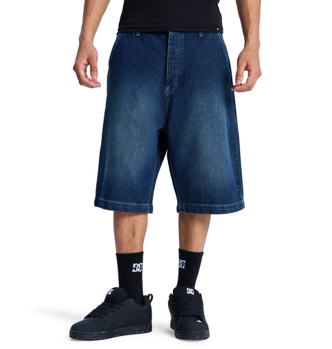 A person wears Reebok Baggy Denim Jorts, black sneakers, a black shirt, and black socks with a white logo. The photo is taken from shoulders down against a white background.