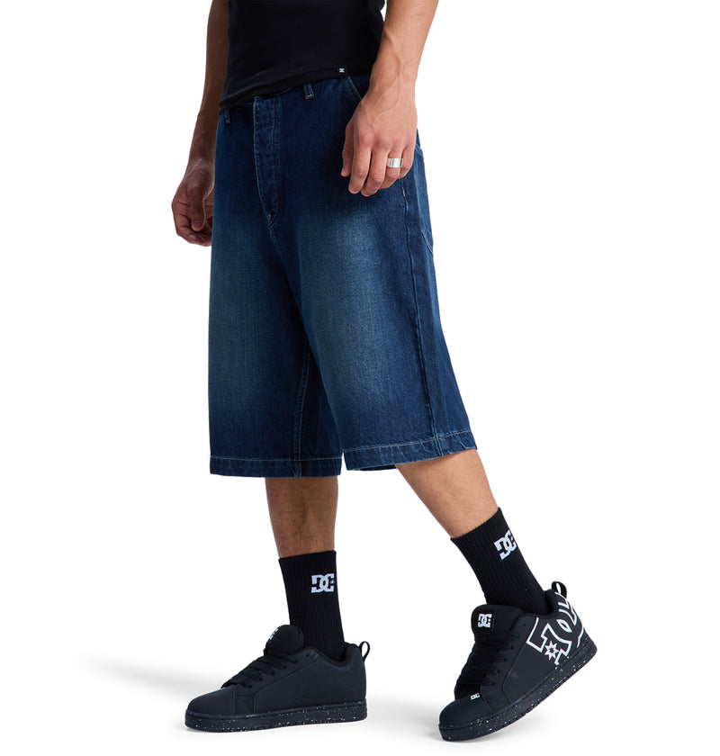 Only the lower half of the body is visible, showing a person wearing Reebok Baggy Denim Jorts that fall below the knee, paired with black socks, black sneakers, and a black t-shirt.