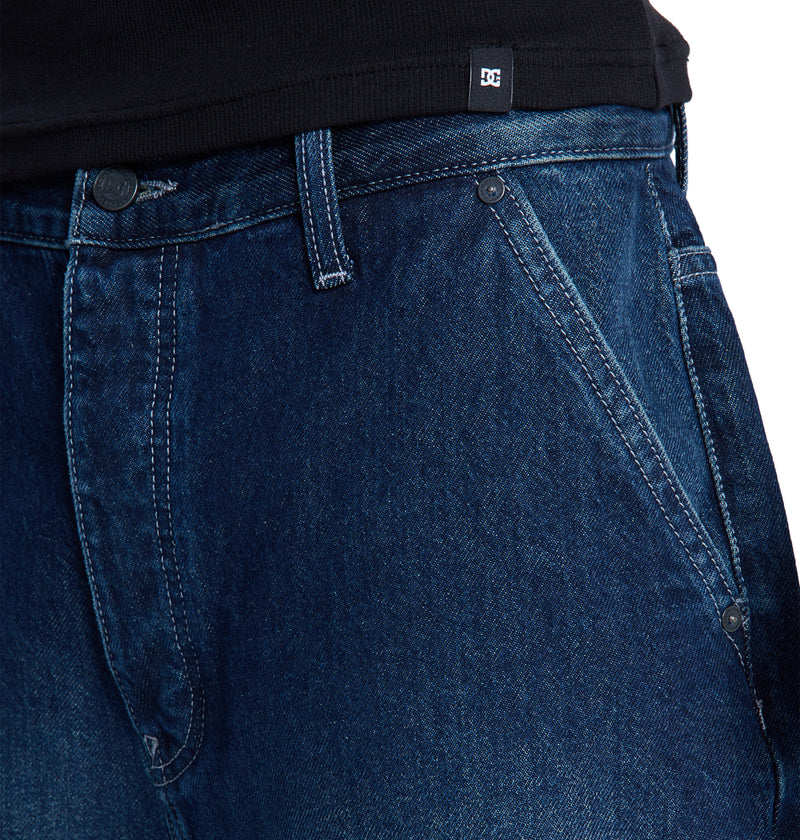 Close-up of a person wearing Reebok's Baggy Denim Jort in dark blue and a black shirt. The jorts feature slanted front pockets, visible stitching, and the waistband alongside the lower part of the shirt are clearly visible.