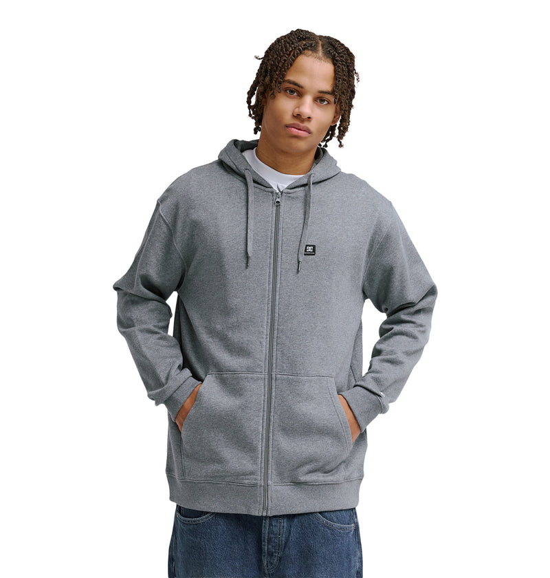 A person in a Reebok Lorion Zh light gray zip-up hoodie with a small logo on the left chest, hands in pockets, stands against a white background. They have short, curly hair and wear blue jeans.