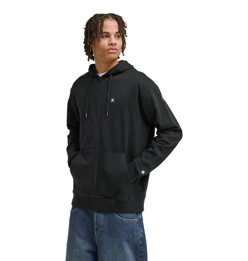 A person with curly hair models the Reebok Lorion Zh black zip-up hoodie with a small white logo on the chest and loose blue jeans, standing against a plain white background.
