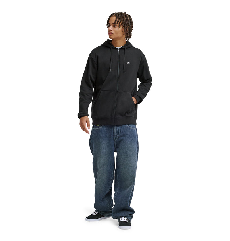 A person with short, curly hair wears a black Reebok Lorion Zh zip-up hoodie, loose blue jeans, and black sneakers while standing against a plain white background.