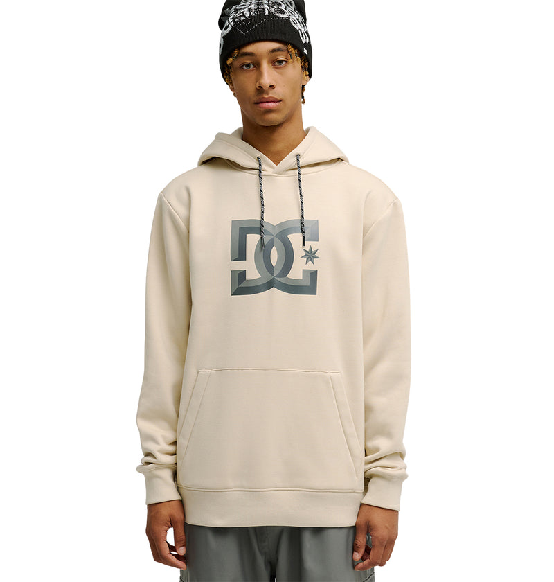 A person wearing the DC Shoes Snowstar beige hoodie, featuring a large DC logo on the front and gray drawstrings, paired with a black and white patterned beanie, stands against a plain white background. The Snowstar hoodie provides a comfortable fit that’s ideal for layering, making it perfect for staying protected on brisk days.