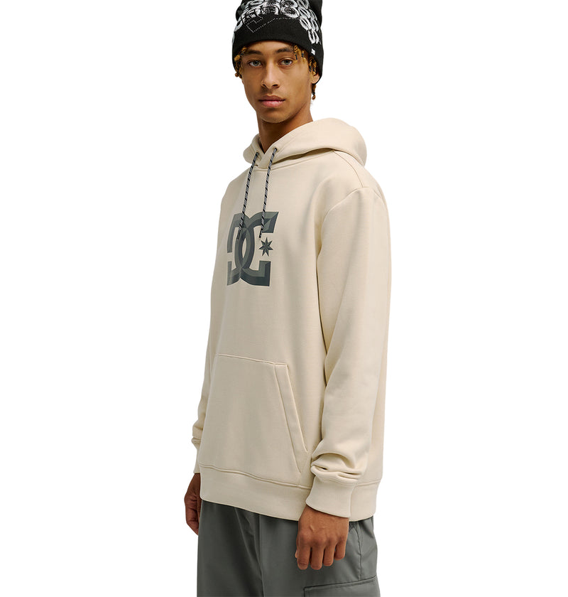 A person wearing the DC Shoes Snowstar beige hoodie, grey shorts, and a black beanie with white designs stands facing slightly to the side against a plain white background, highlighting the Snowstar’s comfortable fit that’s perfect for everyday wear.