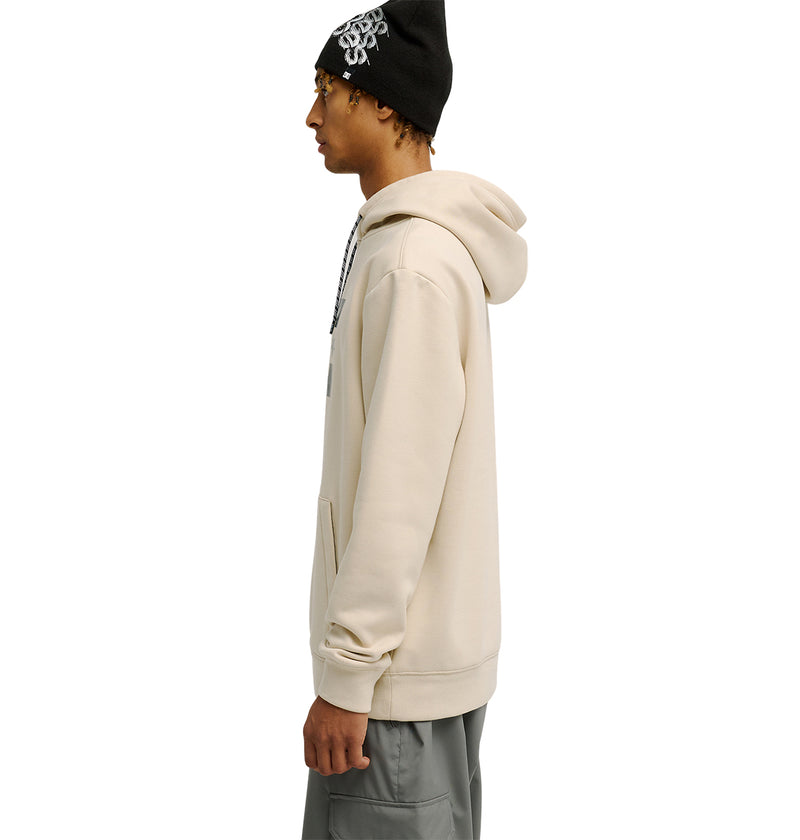 Wearing the DC Shoes Snowstar hoodie in beige, paired with gray pants and a black beanie featuring a white design, a person stands in profile against a white background, highlighting the comfortable fit and weather protection this outfit offers.