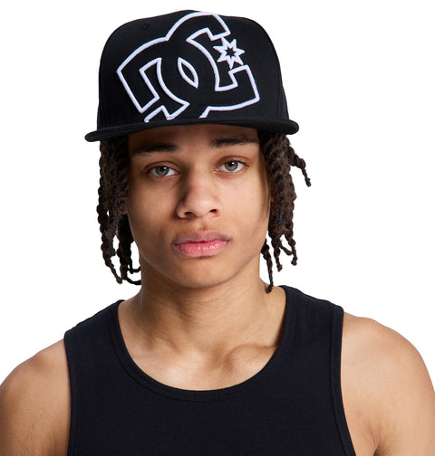 A person with long dreadlocks wears a black Reebok DC Double That Cap and a black tank top, looking directly at the camera against a white background.