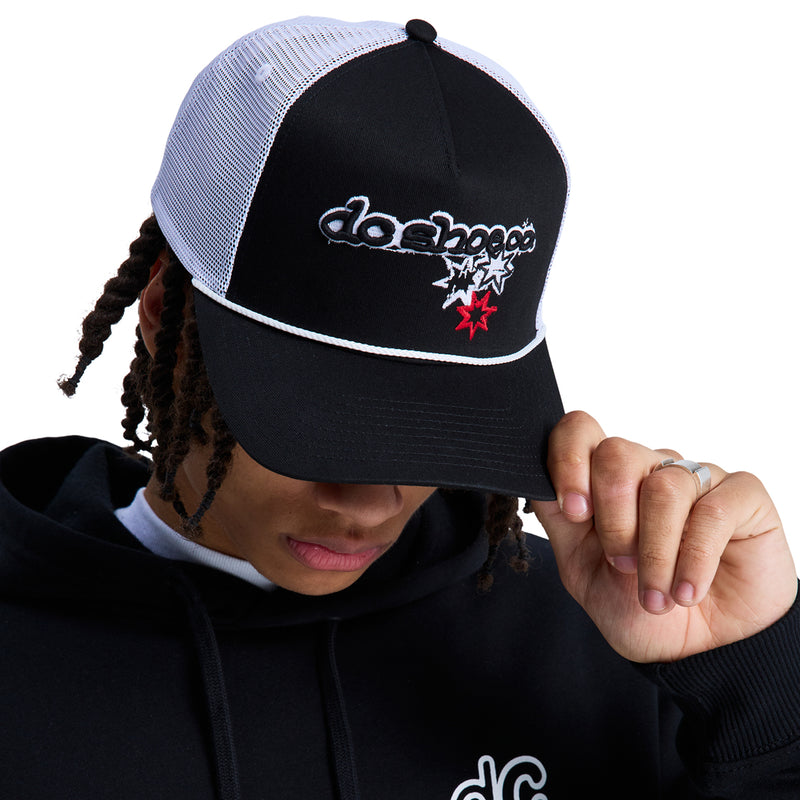 A person tilts the Reebok Side Star Trucker hat—black and white mesh with embroidered stars and text—downward. They wear a black hoodie, have braided hair, and stand against a plain white background.