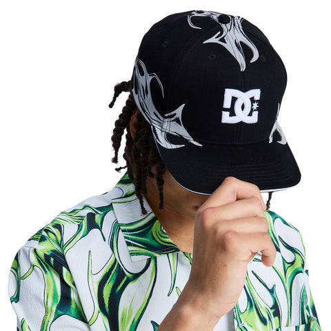 A person wears a black Reebok Fuego Cap with white designs and a green and white patterned shirt, tilting the cap downward to partially cover their face.