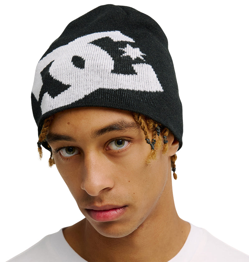 A young person with light brown skin and hazel eyes wears a black Lanai Beanie by DC Shoes, made from recycled fibers and featuring a bold white graphic logo, with short dreadlocks peeking out.