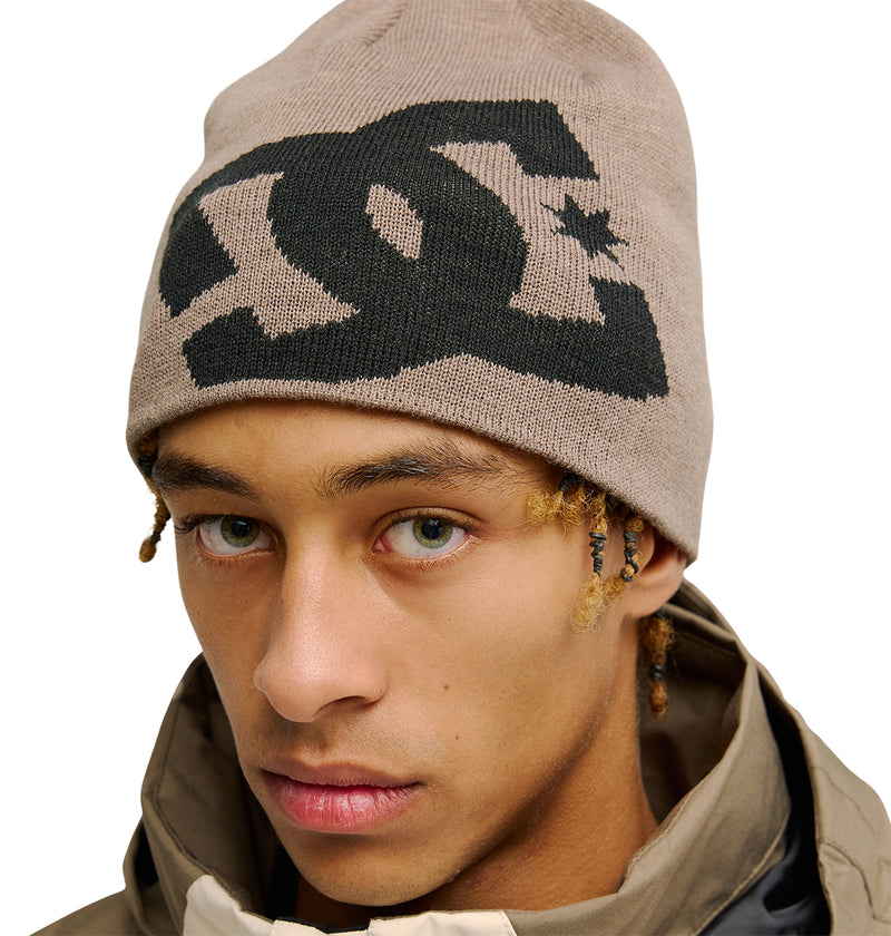 A young person wearing a tan Lanai Beanie by DC Shoes, made from recycled fibers and featuring a bold black logo, pairs it with a brown jacket while looking into the camera with a neutral expression.