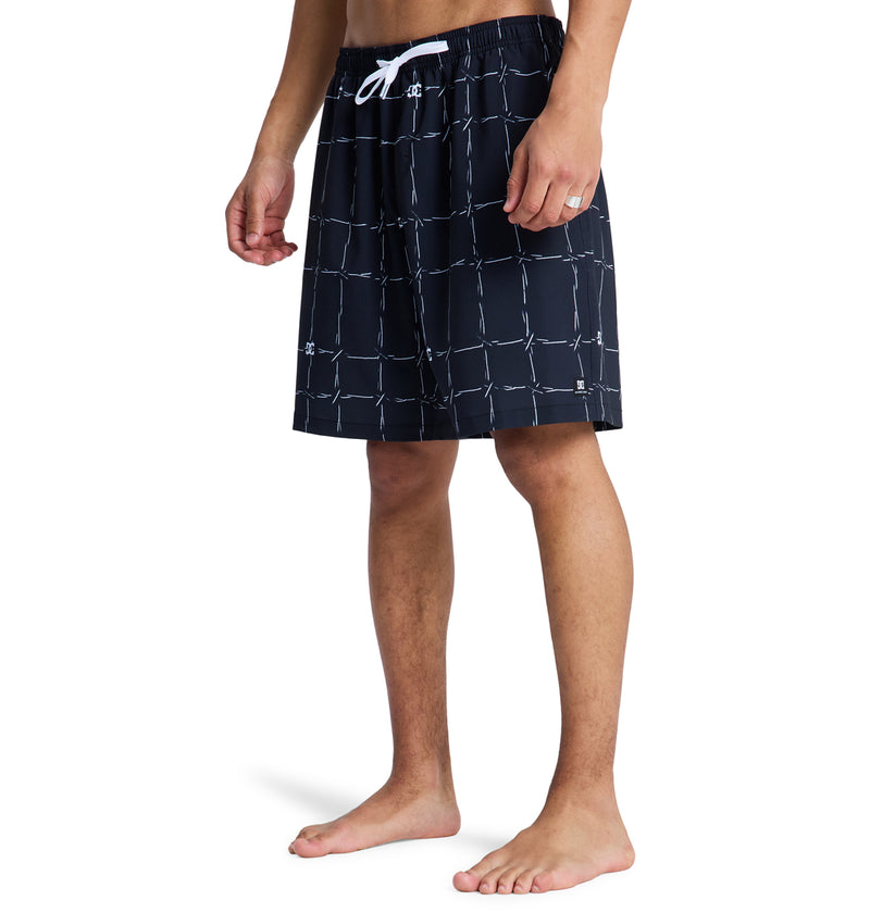 A person wears Reebok DC Wired Volley 19 black swim trunks with a white grid pattern and white drawstring, standing barefoot against a white background. Only their lower body is visible.