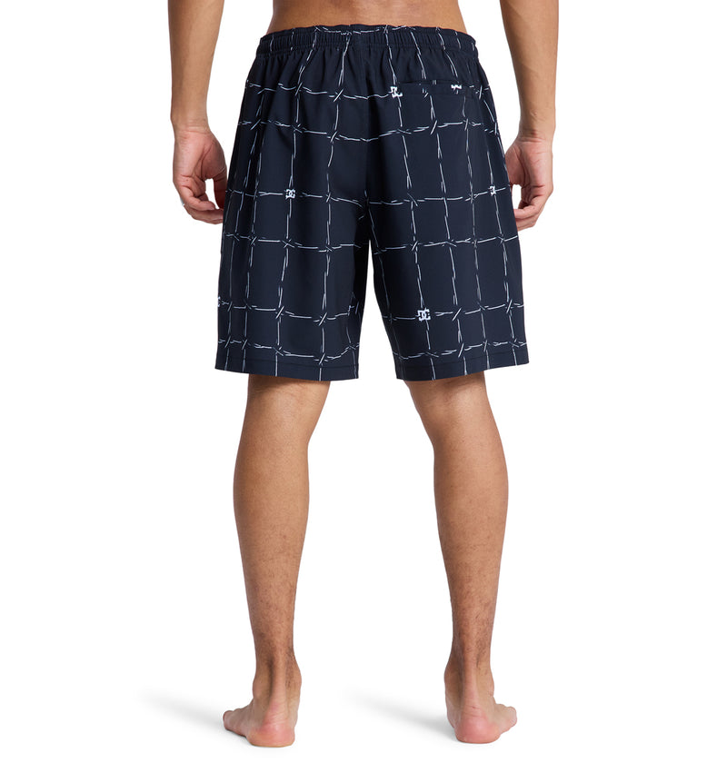 A person stands barefoot with their back to the camera, wearing Reebok DC Wired Volley 19 navy blue shorts with a white grid pattern and an elastic waistband.