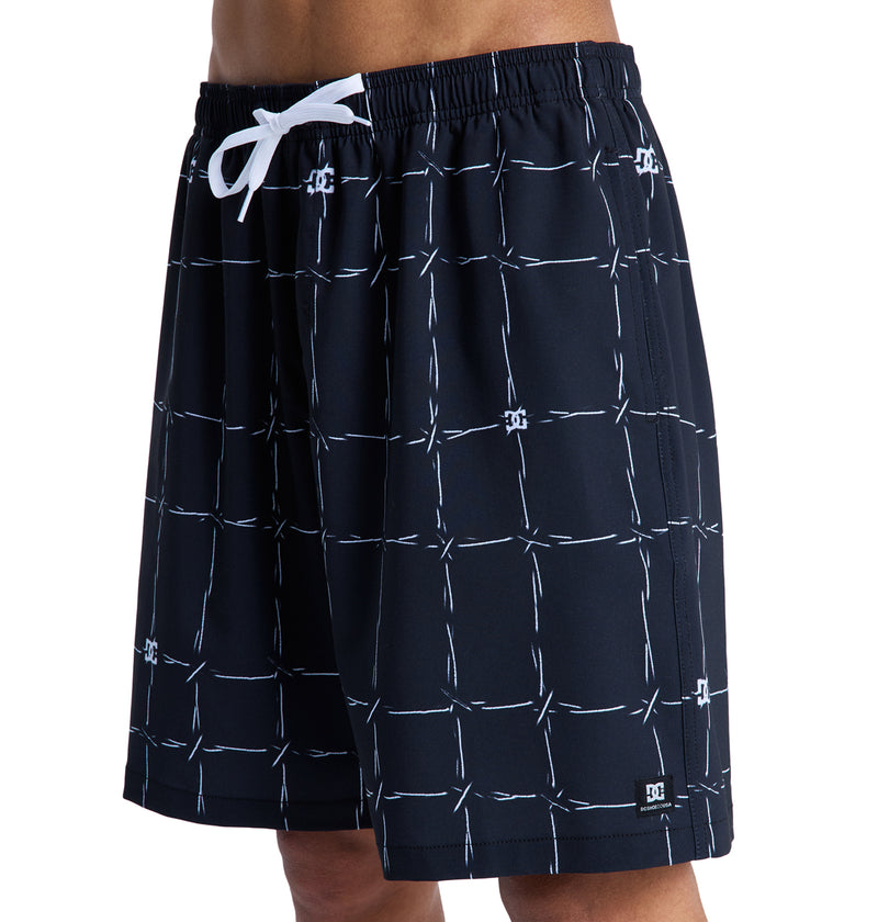 A person wearing Reebok DC Wired Volley 19 black swim trunks with a white grid pattern, white drawstring, and a small DC Shoes logo near the hem.