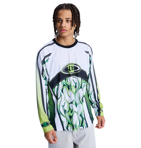 A young man with short dreadlocks models the Reebok Sylem 2 Ls, a long-sleeve white, green, and black graphic shirt with flame patterns, paired with light gray pants against a plain white background.