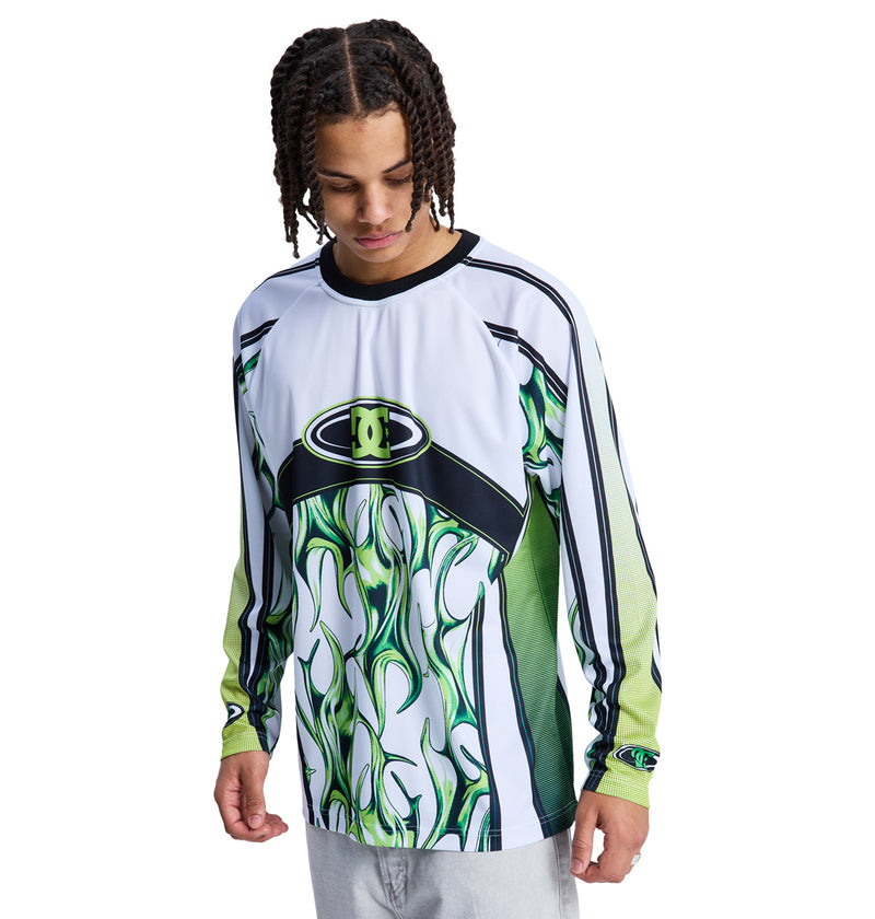 A young man with braided hair looks down while wearing the Reebok Sylem 2 Ls, a colorful long-sleeve shirt featuring green, white, and black flame patterns with an abstract logo, paired with light gray pants against a white background.