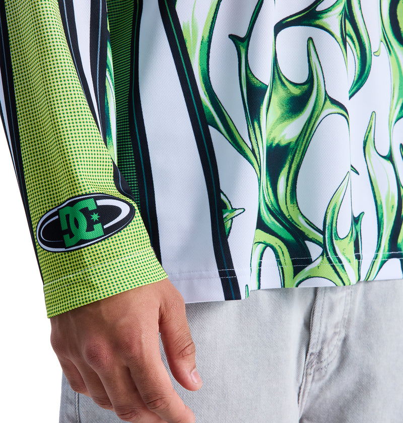 A person wears light denim jeans and a Reebok Sylem 2 Ls long-sleeve shirt featuring green flame graphics and a green-and-black DC logo patch on the wrist. Only the lower half of the shirt and one hand are visible.