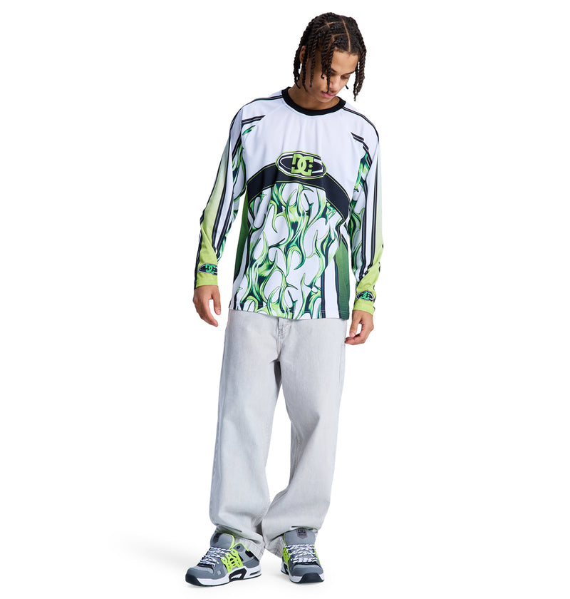 A young man with braided hair wears the Reebok Sylem 2 Ls long-sleeve shirt featuring abstract green and white patterns, paired with loose light gray jeans and gray sneakers with green accents, standing against a white background.